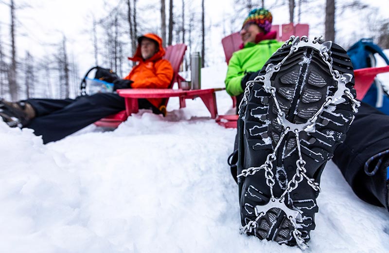 Deux randonneurs sont assis dans des chaises rouges dans la neige. Cadrage serré sur une botte munie d'un crampon.