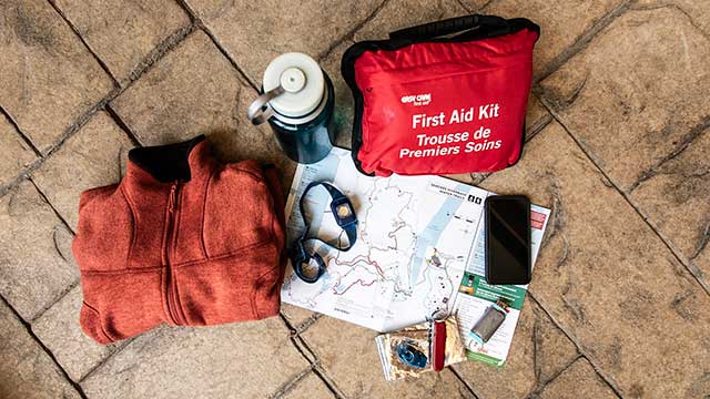 Un chandail, une gourde, une trousse de premiers soins, une carte, un cellulaire, une lampe frontale, un canif, un briquet et un sifflet sont posés sur un plancher.