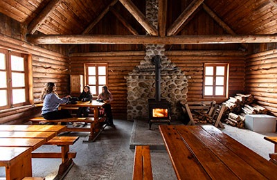 Wooden interior of the Isaïe Shelter, heated with a wood-burning stove, where three people sit at a table.