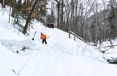 A person shovels snow on a cross-country ski trail in the forest. All-terrain vehicles are parked at the top of a slope on the trail.