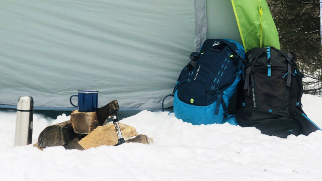 Des sacs à dos, des bûches de bois, une hache, une tasse et un thermos sont posés dans la neige devant une tente.