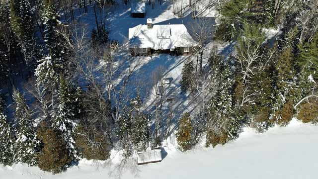 Un gîte du Domaine Wabenaki-Andrew dans une forêt enneigée, devant un lac avec un quai, vus du ciel.