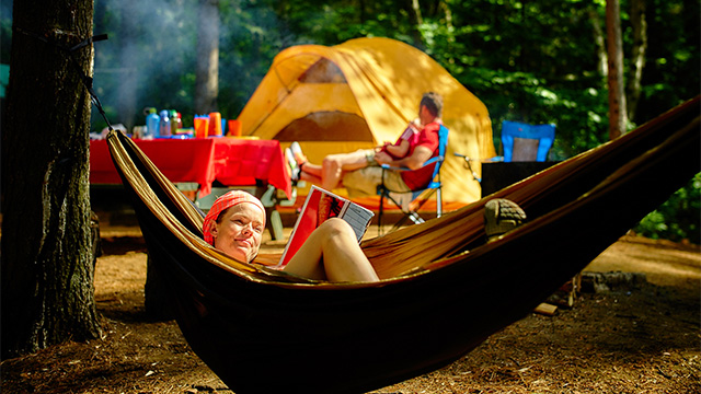 A women calmly reads a book laying in a hamac close to her tent
