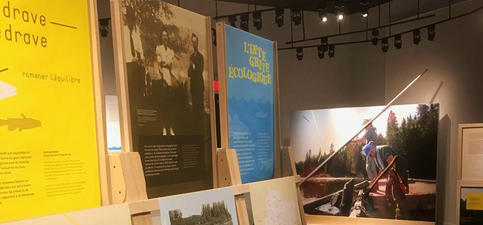 Part of the exhibition of The Restoring Balance: the ups and downs of log-driving exhibition. In the foreground, there are three explanatory panels on the work of the log drive. In the background, there is a huge picture of two log-drivers from the La Mauricie National Park team.

