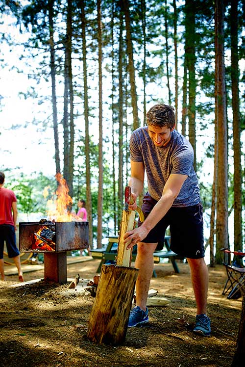 Un jeune homme fend une bûche de bois pour alimenter un feu de camp