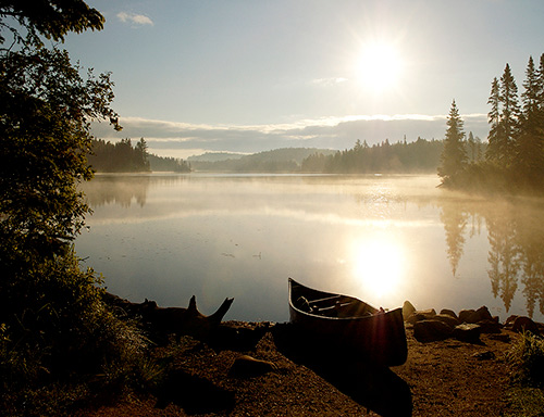 The sun rises on a foggy lake with a canoe on the beach