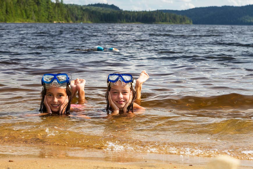 Two smiling girls lie in the water on the shore of a lake. They each wear a diving mask on their heads.