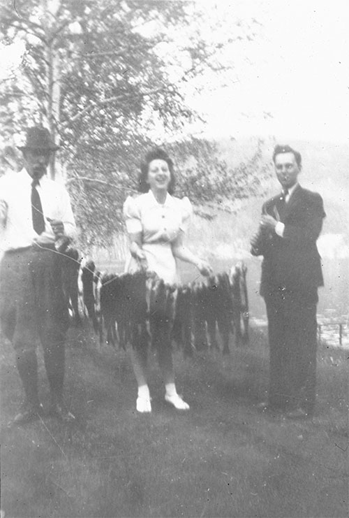 Une femme, accompagnée de deux hommes, tient plusieurs dizaines de poissons dans ses mains au au lac à la Pêche vers 1940