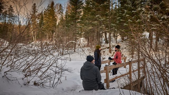 Dans un décor hivernal du parc national de la Mauricie, deux femmes discutent sur un pont de bois enneigé. En avant-plan, un homme assis dans la neige contemple le décor. En arrière-plan, on voit une forêt de conifères.
