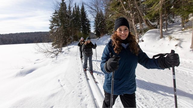 Une mère accompagnée de ses deux filles font du ski de fond, au parc national de la Mauricie, entre une forêt et le lac du Pimbina sous un ciel ensoleillé.