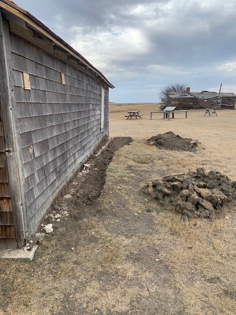 Work was completed on stabilizing the foundations on the Larson homestead buildings. View of the side of a building with disturbed earth at the base of the wall. 
