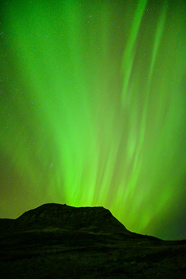 Vibrant green streaks of the Aurora Borealis appear across the night sky as if they are radiating out from a silhouetted hill.