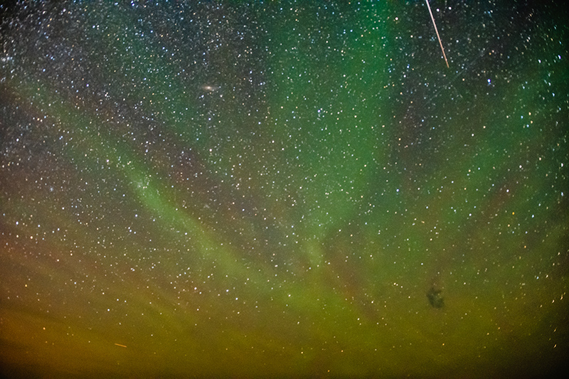 Green, purple, red, and yellow streaks of colour start in the bottom right corner and spread out to all edges of the image against a star filled sky. The white line of a shooting star is visible. 