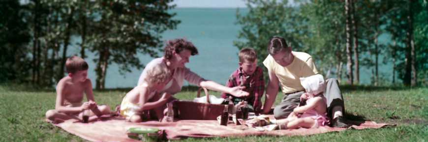 Une famille assise sur une couverture sur une pelouse profite d’un piquenique au bord d’un lac.