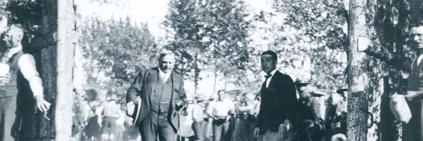 Historical photo of Prime Minister William Lyon McKenzie King in a suit passing through a wooden gate with a crowd of adults. 