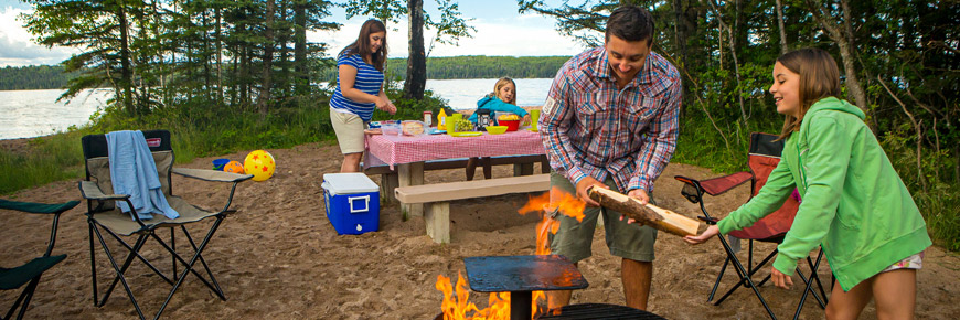 A young girl helps her father add wood to the fire while her sister and mother prepare a picnic lunch. 