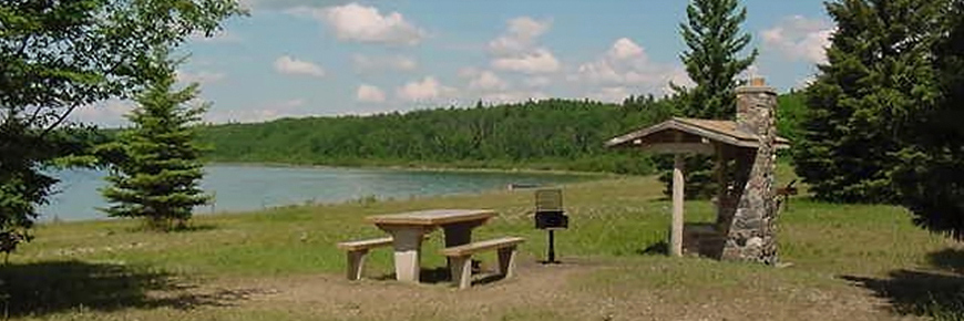 Une image panoramique montre un emplacement de pique nique comportant un foyer en pierre au bord d’un lac.