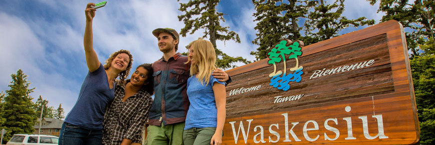 Quatre jeunes adultes prennent une autophoto devant le panneau de bienvenue du village de Waskesiu.