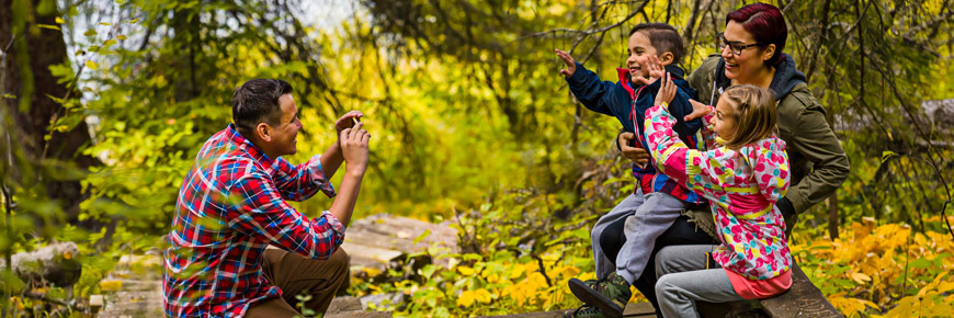 A man takes a photo of his wife and children sitting on a trail-side bench. 
