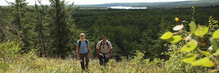 Un jeune couple gravit le flanc d’une colline qui offre une vue panoramique sur un lac et une forêt, derrière.