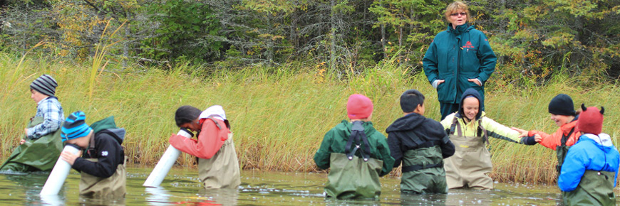 A Parks Canada employee watches as a group of students wade in the river and use equipment to see under the water. 