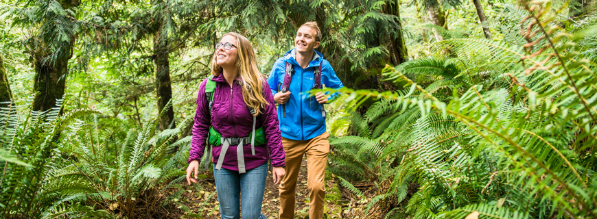 Two people hike along a trail surrounded by dense vegetation.