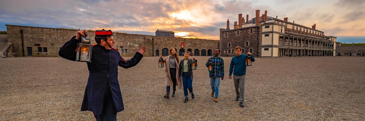 Four visitors and a guide with lanterns walk through the inner courtyard of the Halifax Citadel at dusk.
