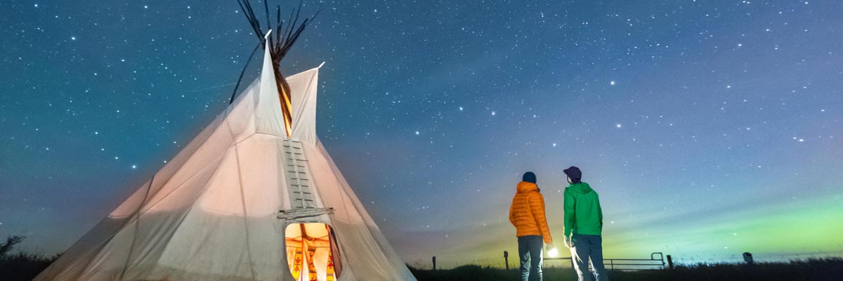 Two visitors gazing at the Big Dipper stars outside a tipi on a starry night at Rocky Mountain House National Historic Site