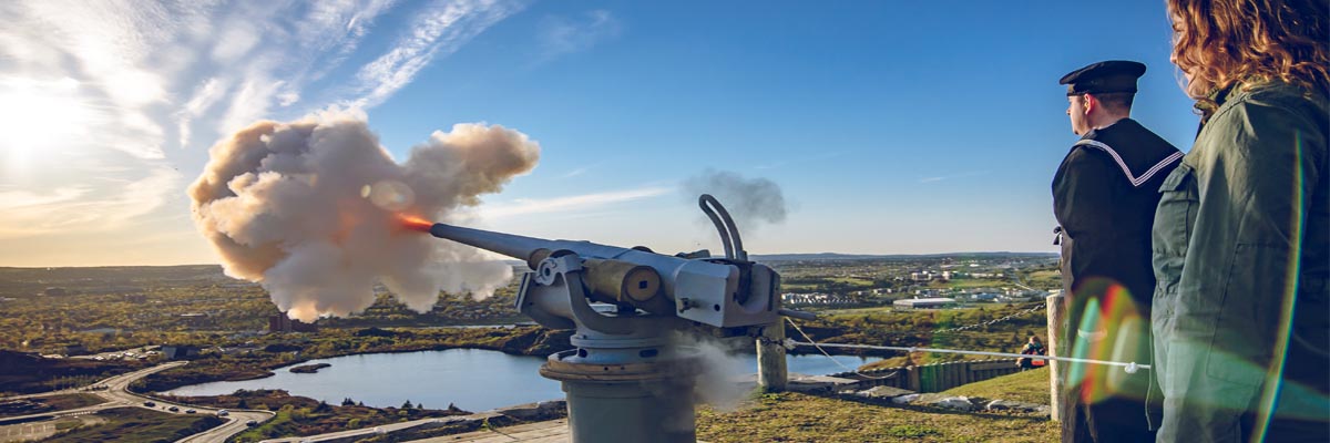 Two adults stand behind a cannon firing on the hillside at Signal Hill National Historic Site.