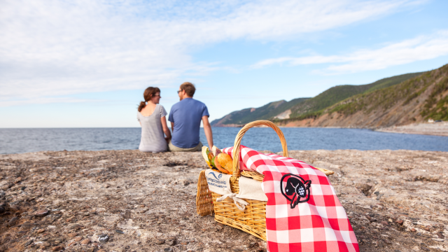 A couple savors in the beautiful view from their perfect picnic place at La Bloc, where they will enjoy their take-out picnic, ordered from a local restaurant, with their Cape Breton Island basket and Parks Canada picnic blanket at Cape Breton Highlands National Park.