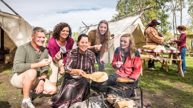 Au camp Métis, une animatrice autochtone fait cuire un pain bannock pour l'offrir à une famille de visiteurs. Un garçon discute de peaux d'animaux avec un animateur Métis au rythme des tambours artisanaux en arrière-plan.