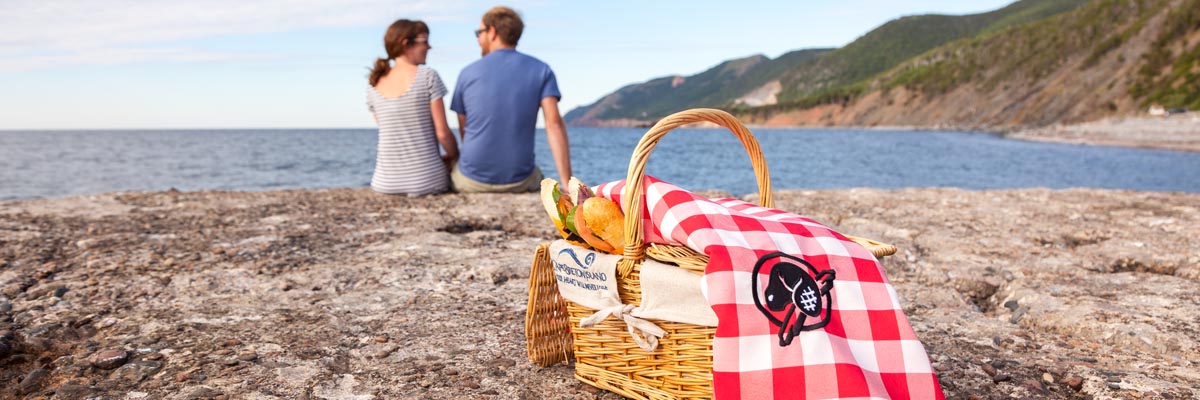 A couple savors in the beautiful view from their perfect picnic place at La Bloc, where they will enjoy their take-out picnic, ordered from a local restaurant, with their Cape Breton Island basket and Parks Canada picnic blanket at Cape Breton Highlands National Park.
