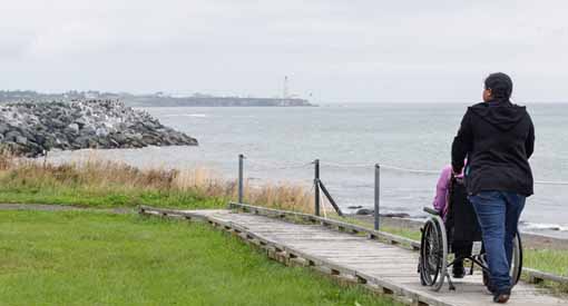 Deux adultes, dont un qui est en fauteuil roulant, se promènent sur un trottoir de bois le long de la mer.