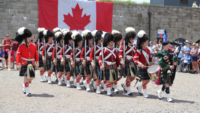 A marching band in traditional costumes march with their instruments with a Canada Flag in the background. 