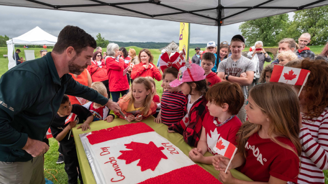 A crowd gathers around a table with a big Canadian flag sheet cake holding mini Canada flags. 