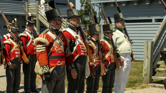 A group of soldiers line up outside a historic fort in costumes holding replica rifle guns. 