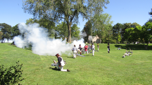Interpreters in soldier unifroms replicate a battle on a grassy field by firing cannons. 