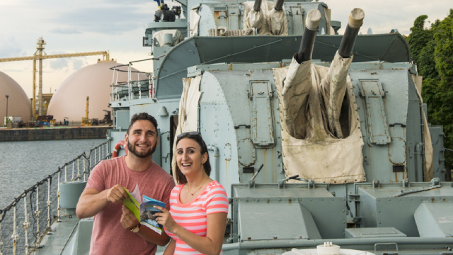 Deux visiteurs avec une brochure sourient devant la caméra à bord du destroyer NCSM Haida.