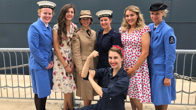 Women in historic costumes from the Second World War pose for a photo in front of the HCMS Haida ship. 