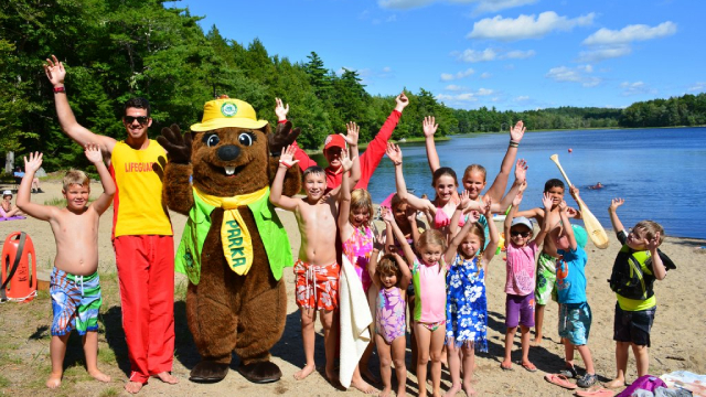 Parka the beaver, Parks Canada's mascot, and a large group of children pose for a photo at the beach with big smiles and thier arms up in the air on a sunny summer day with the lake in the background.