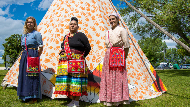 Three women display First Nations beadwork in front of a tipi.