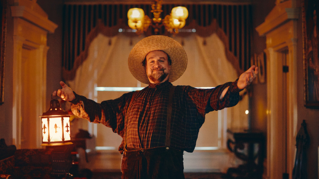 An interpreter wears historic 18th century clothing and stands with his arms out wide while holding a lantern. 