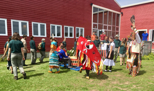 Des membres du personnel, des visiteurs et des guides vêtus de tenues traditionnelles autochtones forment une ronde devant un bâtiment.