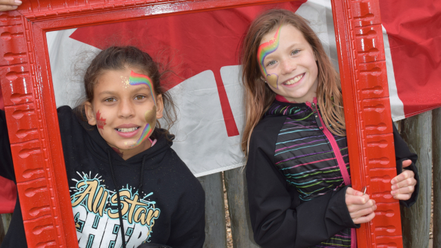 Deux enfants maquillés prennent la pose derrière un cadre rouge avec un drapeau canadien en arrière-plan.