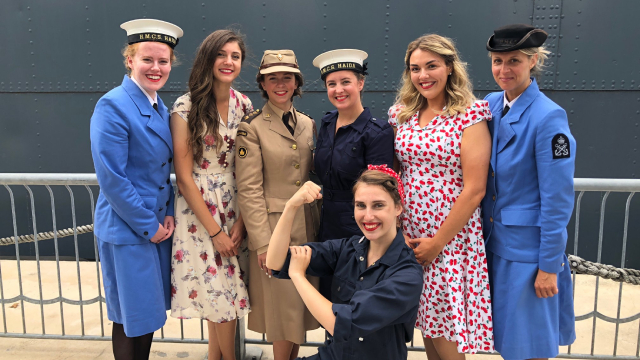 Sept femmes vêtues de différents costumes d’époque posent pour la caméra.