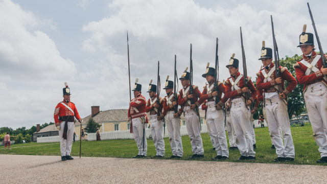 Des personnes en uniforme de soldat, avec un fusil à l’épaule, sont alignées en rang devant des bâtiments historiques.