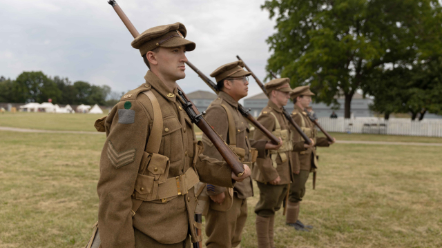Des acteurs de reconstitution en uniforme de soldat, avec un fusil à l’épaule, sont alignés en rang devant des bâtiments historiques.