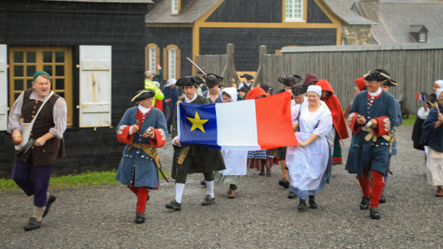 Des guides en costume d’époque portant un drapeau acadien marchent devant des bâtiments historiques.