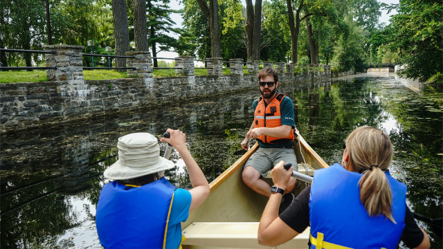 Un guide de Parcs Canada et des visiteurs en rabaska sur un canal bordé d’arbres.