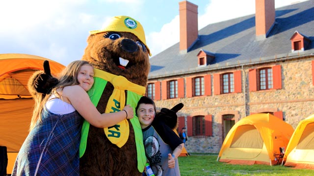 Deux jeunes enfants posent avec la mascotte Parka avec des tentes et un bâtiment historique en arrière-plan.